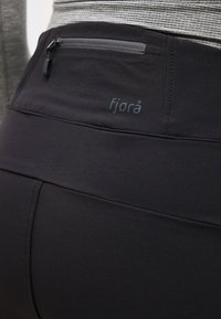 Detailný záber osoby nosiacej čierne legíny značky Fjora s vreckom na zips na chrbte a sivý pruhovaný top.