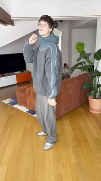 Obszerna szara kurtka polarowa z turkusowymi akcentami i wysokim kołnierzem, zestawiona z luźnymi szarymi spodniami i srebrnymi sneakersami. Wnętrze pomieszczenia.