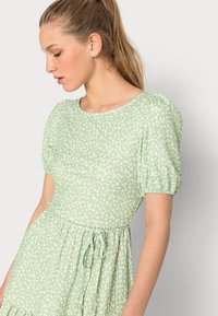 Vestido verde claro feito de tecido texturizado com padrões florais brancos. Apresenta mangas bufantes e um cós com cordão para um ajuste suave.