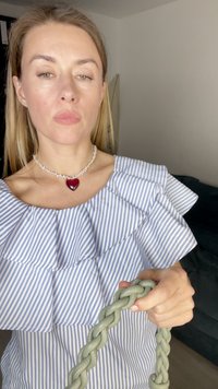 Sininen ja valkoinen raidallinen pusero, jossa on röyhelökaulus, yhdistettynä sydämenmuotoiseen punais-valkoiseen helmikaulaketjuun, pitäen kädessään letitettyä vihreää käsilaukkua.