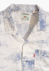 Chemise Levi's blanche à coupe décontractée avec imprimé paysage désertique bleu, boutonnée devant, col, poche poitrine et étiquette Levi's à l'intérieur du col et sur la poche.