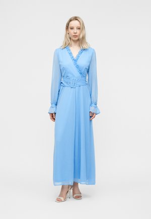 Femme debout portant une robe portefeuille bleu clair à manches longues avec bordure à volants, large ceinture et talons blancs ouverts sur un fond uni.