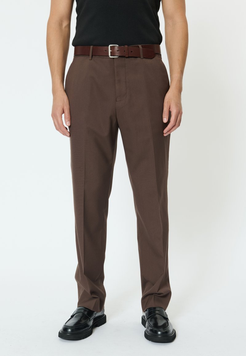 Mand iført brune skræddersyede bukser, en sort indstikket skjorte, et brunt læderbælte og sorte lædersko, stående mod en hvid baggrund.