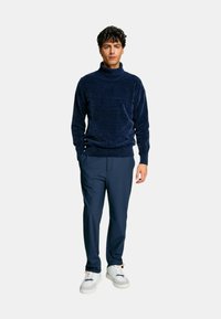 Maglione a collo alto blu navy con un tessuto a trama, abbinato a pantaloni blu scuro e sneakers bianche con dettagli arancioni.