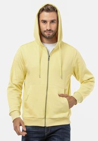 Geel ritsjack met een voorzak, trekkoorden, ribgebreide manchetten en zoom. Zacht materiaal met een casual ontwerp.