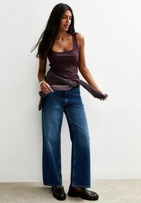 Vrouw met lang donker haar, gekleed in een mouwloze paarse top, wijde blauwe jeans, zwarte instappers en een trui om haar middel geknoopt.