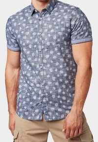 Camicia a maniche corte con bottoni in blu, caratterizzata da un motivo testurizzato di forme bianche. Presenta un colletto classico e maniche arrotolate.