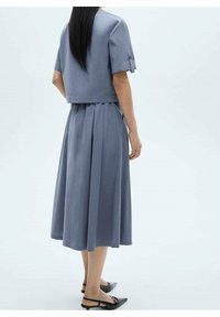 Donna con lunghi capelli neri, che indossa una maglietta blu a maniche corte e una gonna midi plissettata coordinata, con scarpe slingback a punta nera.
