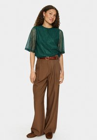 Blouse en dentelle teal à manches courtes, associée à un pantalon large marron et une ceinture marron, portée avec des mocassins marron. Motifs texturés subtils.