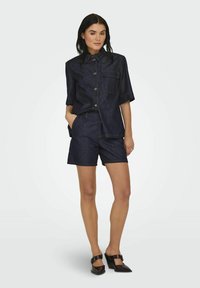 Tumši džinsa pogu krekls ar īsām piedurknēm un kabatu, kombinēts ar atbilstošām tumši džinsa šortiem. Valkāts ar melniem augstpapēžu mūlēm.