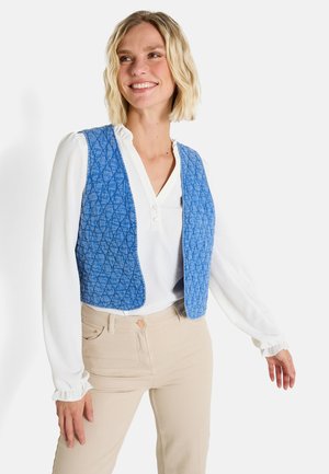 Chaleco acolchado azul con patrón de diamantes, de frente abierto, superpuesto sobre una blusa blanca con cuello arruga, combinado con pantalones beige.