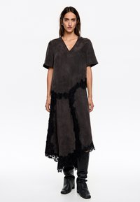 Vestido de ante gris con escote en V, de corte suelto, con detalles de encaje negro a lo largo del dobladillo y los lados, y mangas cortas. Combinado con botas negras.