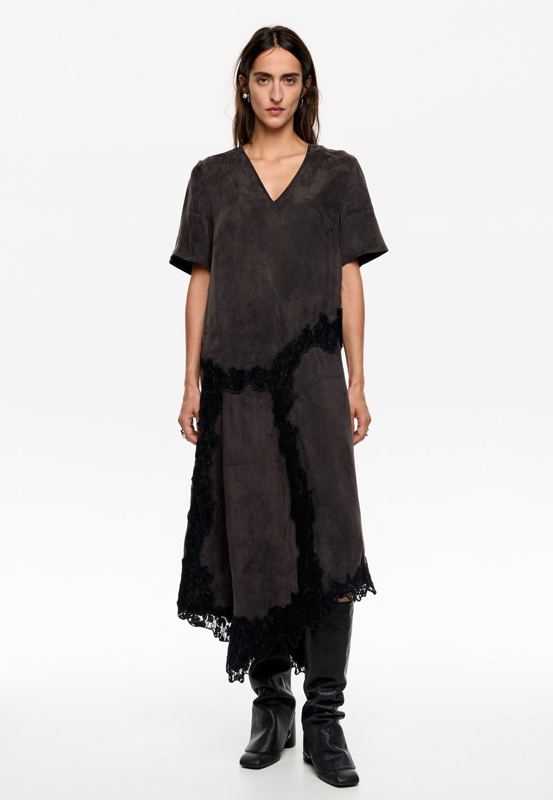 Vestido de ante gris con escote en V, de corte suelto, con detalles de encaje negro a lo largo del dobladillo y los lados, y mangas cortas. Combinado con botas negras.