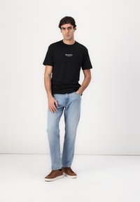 Sort bomulds t-shirt med korte ærmer og hvid tekst på fronten, parret med lyseblå jeans og brune sneakers, stående foran en hvid baggrund.