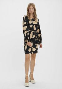 Black dress with large beige abstract patterns, long sleeves, and a waist tie. Smooth fabric, knee-length, and paired with chunky white heels.