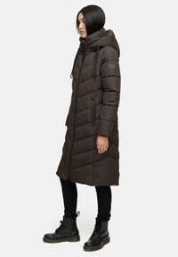 Cappotto puffer marrone con cappuccio, caratterizzato da un design trapuntato e chiusura frontale con zip. Abbinato a jeans slim neri e stivali neri con lacci.