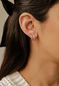 Srebrne kolczyki w formie podwójnych obręczy, odbijające światło dzięki gładkiej teksturze powierzchni, noszone na uchu z ciemnymi włosami w pobliżu.