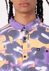 Cămașă cu mânecă scurtă, având un model abstract vibrant în nuanțe de violet, galben și gri. Fabricată dintr-un material ușor, cu închidere cu nasturi și buzunar.