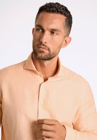 Helles Pfirsichfarbendes Leinenhemd mit Button-Down-Kragen, sichtbaren Knöpfen und einer entspannten Passform. Die Textur wirkt weich und glatt.