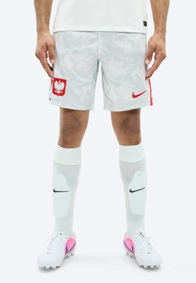 Πόδια που φορούν άσπρες και γκρι σορτς ποδοσφαίρου της Πολωνίας, λευκές επιγονατίδες Nike και λευκά παπούτσια ποδοσφαίρου με ροζ και μοβ λεπτομέρειες.