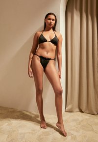 Biquíni preto com copas triangulares e laterais ajustáveis, apresentando um tecido suave e um design minimalista. A modelo está de pé sobre um chão texturizado.