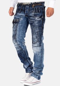 Jeans cargo in denim blu con molteplici tasche, dettagli sbiaditi e un accento con cerniera. Abbinati a sneakers bianche con lacci.