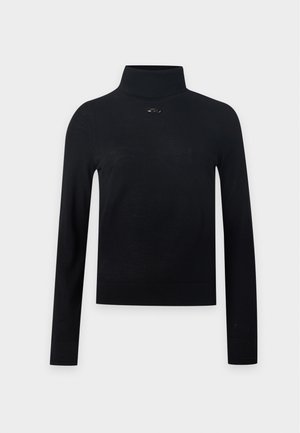 Femme portant un col roulé noir à manches longues, un short sombre jusqu'aux genoux, une ceinture blanche, des sandales à talons noires et tenant un petit sac à main noir.