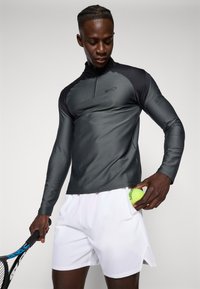 Haut de sport à manches longues en gris foncé avec un tissu texturé, doté d'une demi-fermeture éclair et d'accents noirs contrastés, porté avec un short blanc et tenant une balle de tennis.