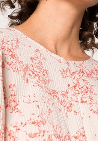 Naisten kaula ja ylempi rinta, joissa on päällä teksturoitu valkoinen paita punaisilla kukkakuviolla ja pieni vetoketju kaula-aukossa.