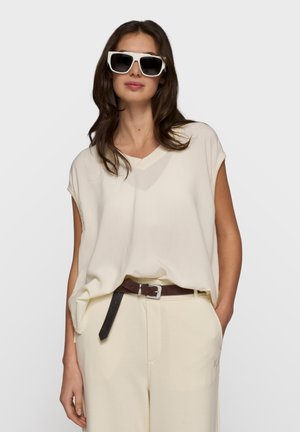 Kobieta nosząca duże białe okulary przeciwsłoneczne, luźny kremowy top bez rękawów, kremowe spodnie i brązowy pasek, z jedną ręką w kieszeni.