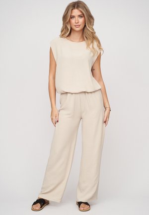 Femme aux longs cheveux blonds portant un haut beige ample sans manches et un pantalon large assorti avec des sandales noires.