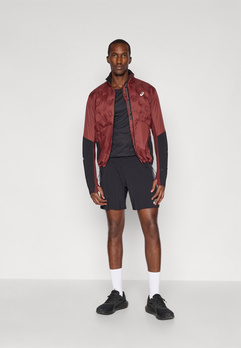 Veste matelassée bordeaux avec des manches longues noires, portée sur un t-shirt à manches courtes noir et un short noir, assortie de chaussettes blanches et de baskets noires.