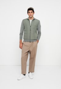 Man wearing a green quilted vest over a gray long-sleeve shirt, beige wide-legged pants, white sneakers, and a plaid cap standing with hands in pockets.