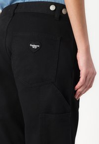 Black canvas pants with a heart-shaped Carhartt WIP patch on the back pocket. Features two metal buttons at the waist and diagonal pocket design.