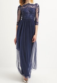 Nocna sukienka w kolorze granatowym z dopasowanym gorsetem ozdobionym koronką i przejrzystymi rękawami o długości trzech czwartej, falującą spódnicą z tiulu oraz eleganckimi srebrnymi szpilkami.