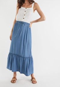 Débardeur en lin blanc avec boutons sur le devant, associé à une longue jupe en denim bleu avec un ourlet à volants froncés et une taille à cordon. Sandales marron.