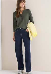 Femme portant un haut henley vert à manches longues, un jean bleu foncé, des baskets argentées, tenant un pull jaune en tricot plié, souriante, debout contre un mur beige.