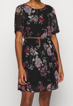 Woman wearing a black floral dress with short sleeves and a braided brown belt at the waist against a plain background.