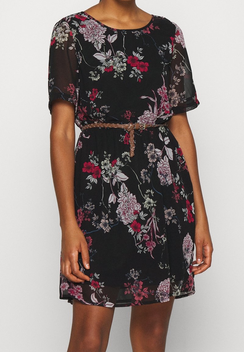 Femme portant une robe noire à fleurs avec des manches courtes et une ceinture tressée marron à la taille devant un fond uni.