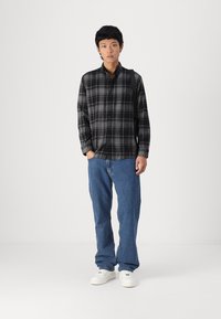 Μαύρο και γκρι καρό πουκάμισο με μακριά μανίκια, συνδυασμένο με μπλε τζιν παντελόνι και λευκά αθλητικά παπούτσια, σε απλή φόντο.