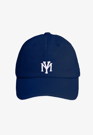 Jonge man met een marineblauwe pet met beige NY-logo en een marineblauw shirt met rode en blauwe afbeelding, kijkt omlaag tegen een effen achtergrond.