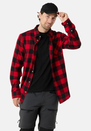 Chemise en flanelle à carreaux rouges et noirs, à manches longues, avec un devant boutonné, portée par-dessus un tee-shirt noir, associée à un short foncé et une casquette noire.