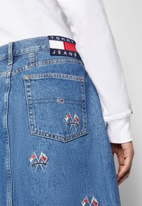 Nahaufnahme einer Person, die blaue Jeans aus Denim mit gestickten kleinen gekreuzten Flaggen und einem Tommy Jeans-Label trägt, kombiniert mit einem weißen Langarmshirt.