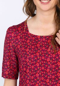 Bluse mit Blumenmuster, die einen quadratischen Ausschnitt und kurze Ärmel hat. Der Stoff ist überwiegend dunkel lila mit kleinen roten und grünen Blumen.