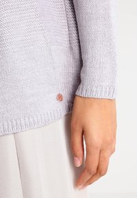 Pull en maille gris clair avec un motif texturé, poignets côtelés et un petit logo métallique sur le bas.