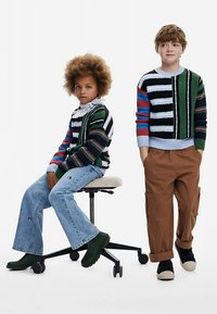 Flerfärgade stickade tröjor med ränder, jeans och vardagliga skor. Ett barn sitter på en pall; det andra står bredvid.