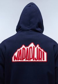 Mörkblå huvtröja med en stor, vit och röd broderad logotyp med bergsformer på baksidan. Tillverkad av bommaterial med mjuk textur.