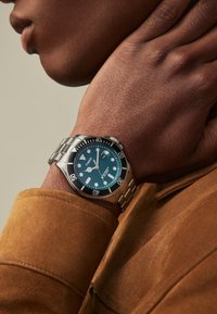 Reloj de acero inoxidable plateado con una esfera verde oscuro, marcadores de hora luminosos, bisel negro y pulsera metálica en una muñeca contra un fondo de tela marrón.