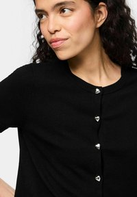 Musta neulecardigan, jossa on pyöreä pääntie, lyhyet hihat ja sydämenmuotoiset hopeanväriset napit edessä. Pehmeä neulottu materiaali, jolla on sileä pinta.