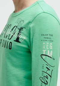 Langärmliges grünes T-Shirt aus weichem Stoff, mit schwarzem Druck und Text sowie Akzenten auf der Vorderseite und dem rechten Ärmel.